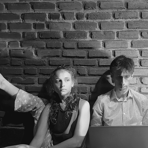 Uma mulher com a perna ao lado e um homem num computador em frente a uma parede de tijolos